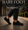 A man running down a tree-lined path wearing a black athletic t-shirt, black shorts, and black running shoes, with another person's legs in black jeans and black shoes in the foreground. Large text overlay reads 'BAREFOOT by loungers'.