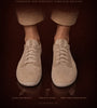 A pair of beige knit lounge sneakers shown on a person's bare feet, with rolled-up tan pants visible against a dark brown background. Large text at the top reads 'LOUNGERS' and smaller text includes 'HANDMADE FOR BAREFOOT CONCIUS WALKING' and feature callouts near the bottom.